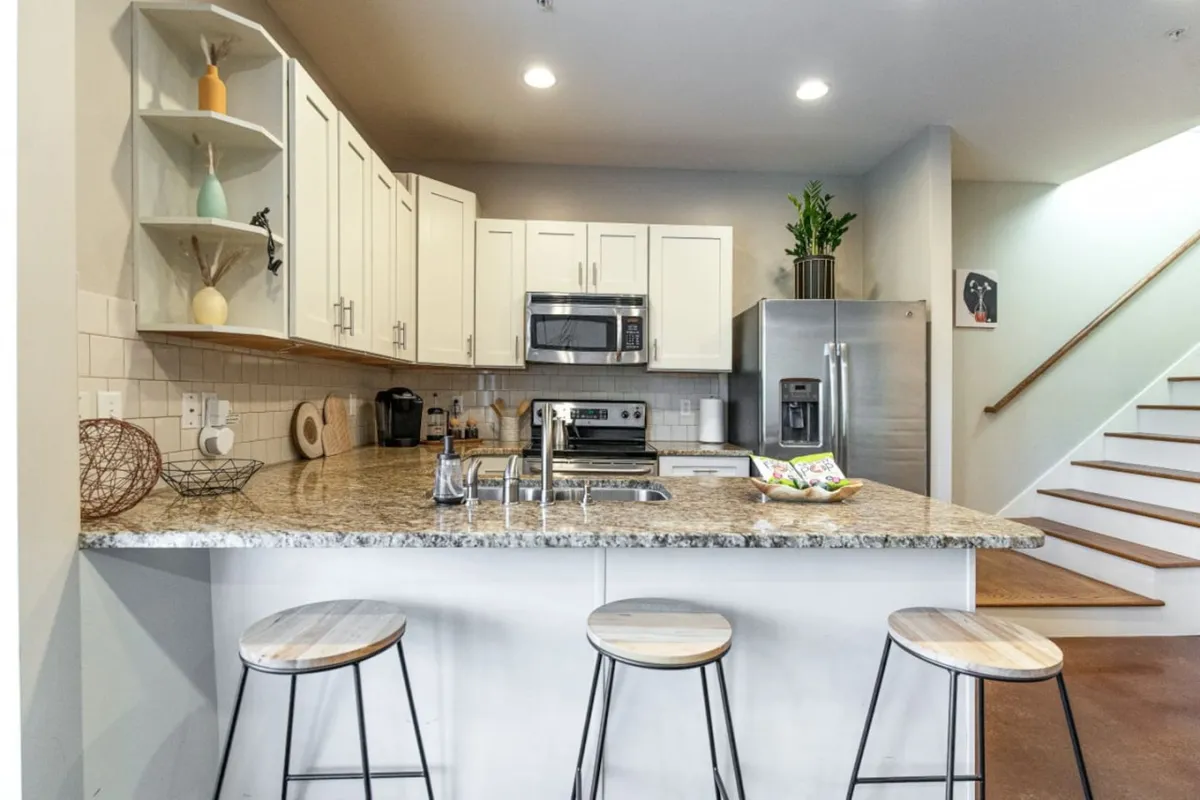 A bright, modern kitchen featuring a granite countertop, gleaming stainless steel appliances, open shelving with decorative accents, and stools at a spacious island. The scene radiates an inviting, functional atmosphere, perfect for meal prep or gathering around for coffee and conversation.