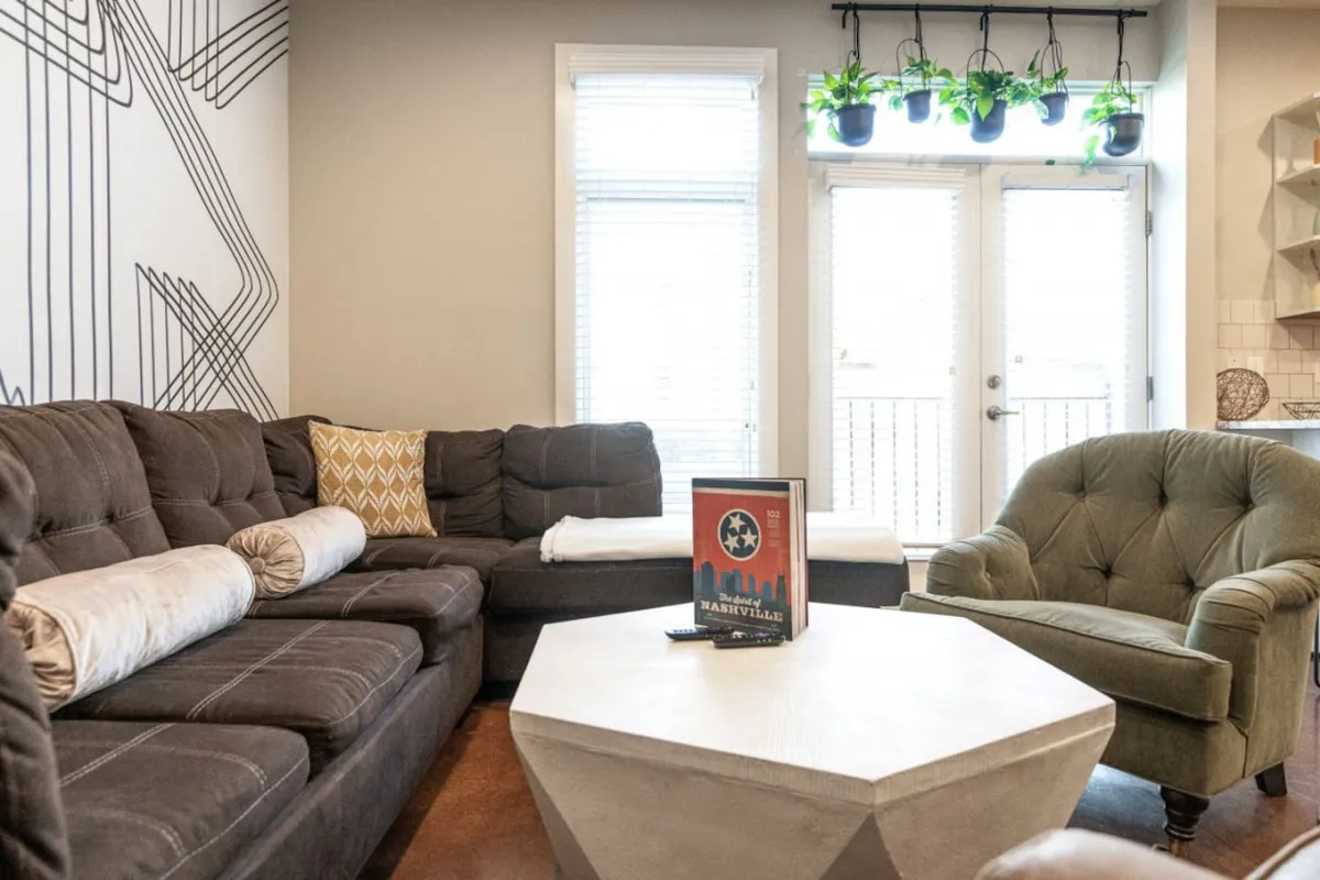 A modern, cozy living area with a large sectional sofa, geometric wall art, unique coffee table, and lush house plants. The space is designed for relaxing, entertaining guests, or unwinding with a good book.