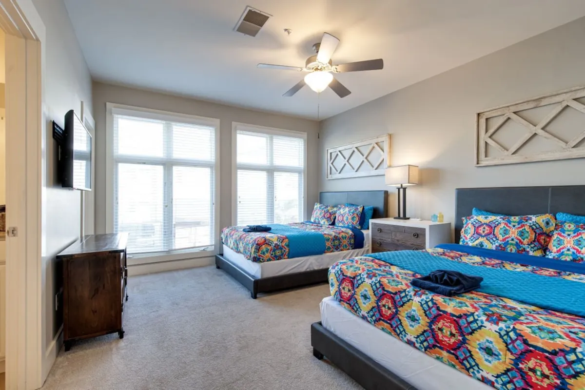 A bright, cheerful bedroom with colorful bedding, large windows letting in natural light, and a nightstand with a lamp. The space feels welcoming and comfortable, suggesting a relaxing retreat perfect for families after a day of exploring Nashville.