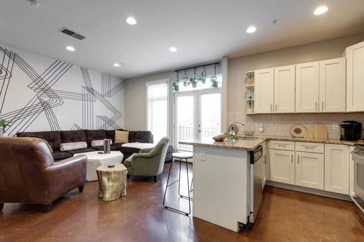 This image features a stylish living room with a comfortable sofa, kitchen island, artistic wall art, and large windows. The room invites socializing, cooking, and relaxing together, aligning with the section's focus on gathering spaces.