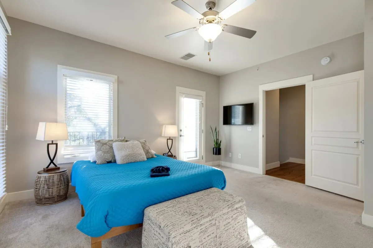 This image depicts a serene bedroom featuring a blue bedspread, decorative throw pillows, and nightstands with lamps flanking the bed. A wall-mounted TV and a window with blinds are visible, offering both entertainment and natural light. The overall ambiance is calm and cozy, ideal for relaxation or watching a show in bed.